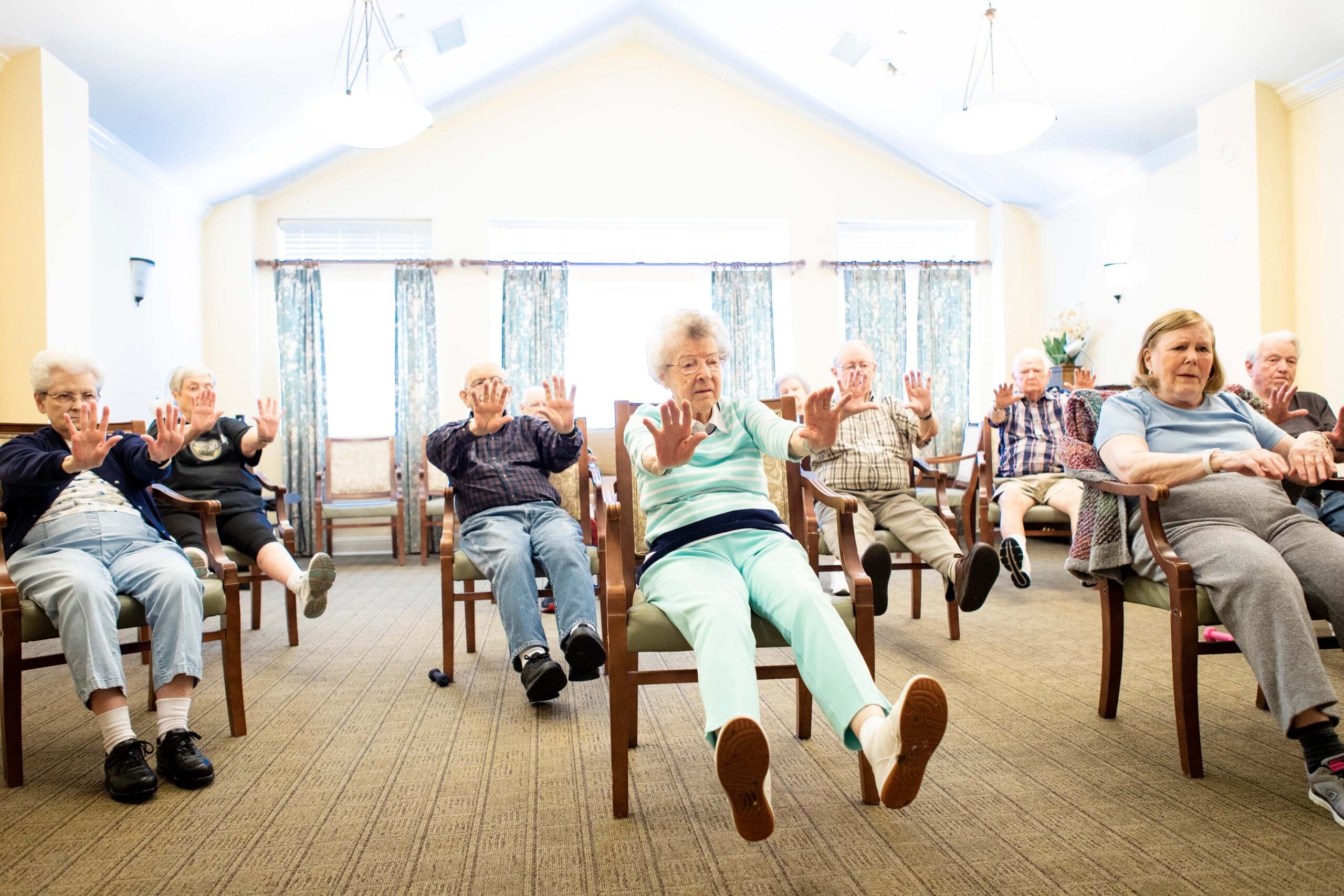 A group of white seniors are sitting on chairs that are spread our across the room, stretching their arms and feet forward.