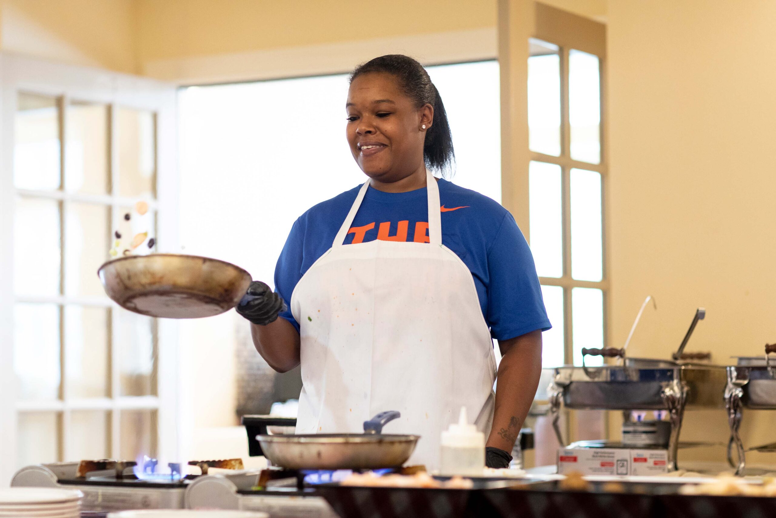 Grace_Management_Memphis_2018-5857 - Palm Bay A middle-aged black woman wearing a white apron is standing in front of the stove with a frying pan in her hand, tossing vegetables up in the air.